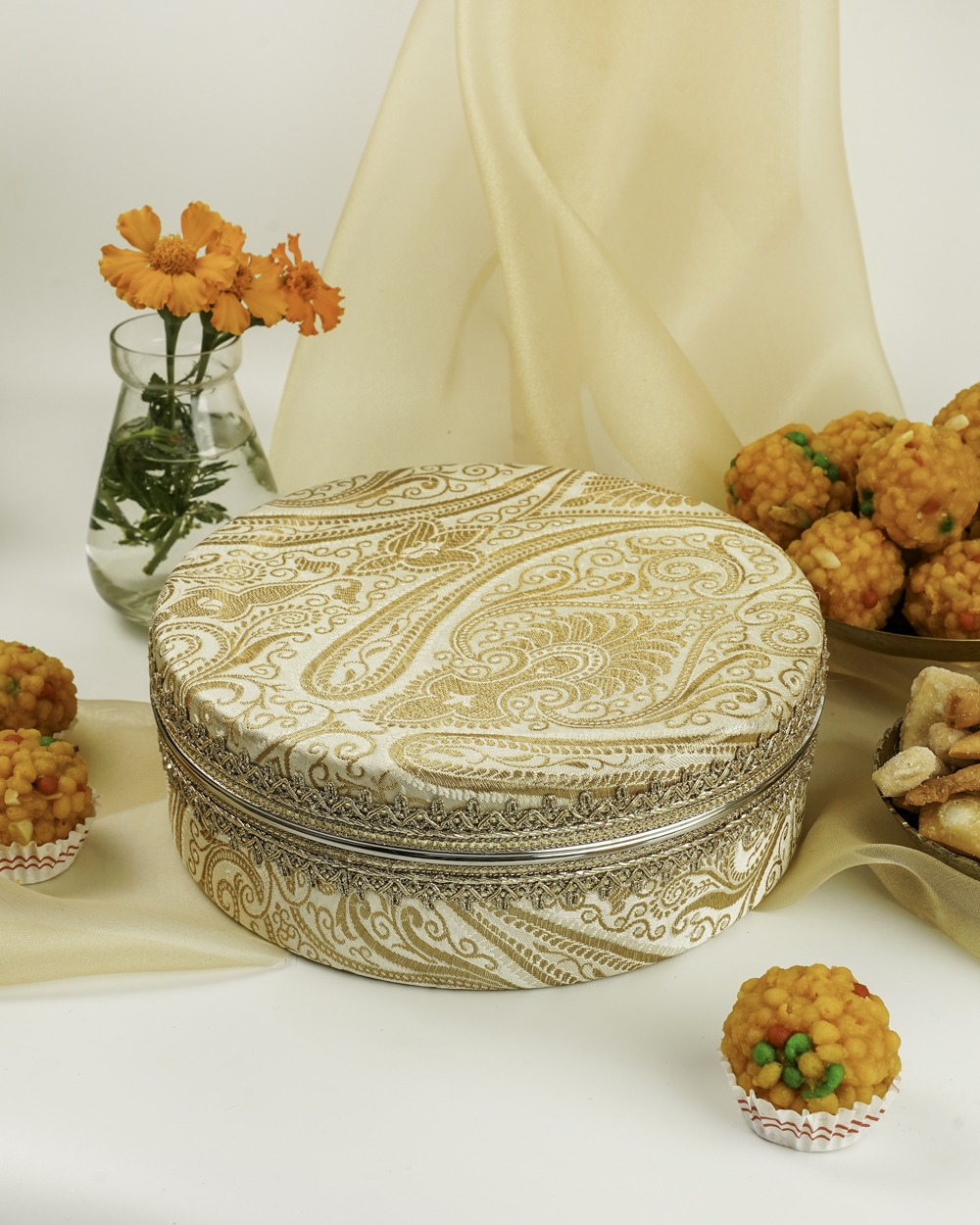 Elegant cream Noori Ladoo Box by Navka Studio with gold paisley brocade and silver lace trim, presented with festive sweets.