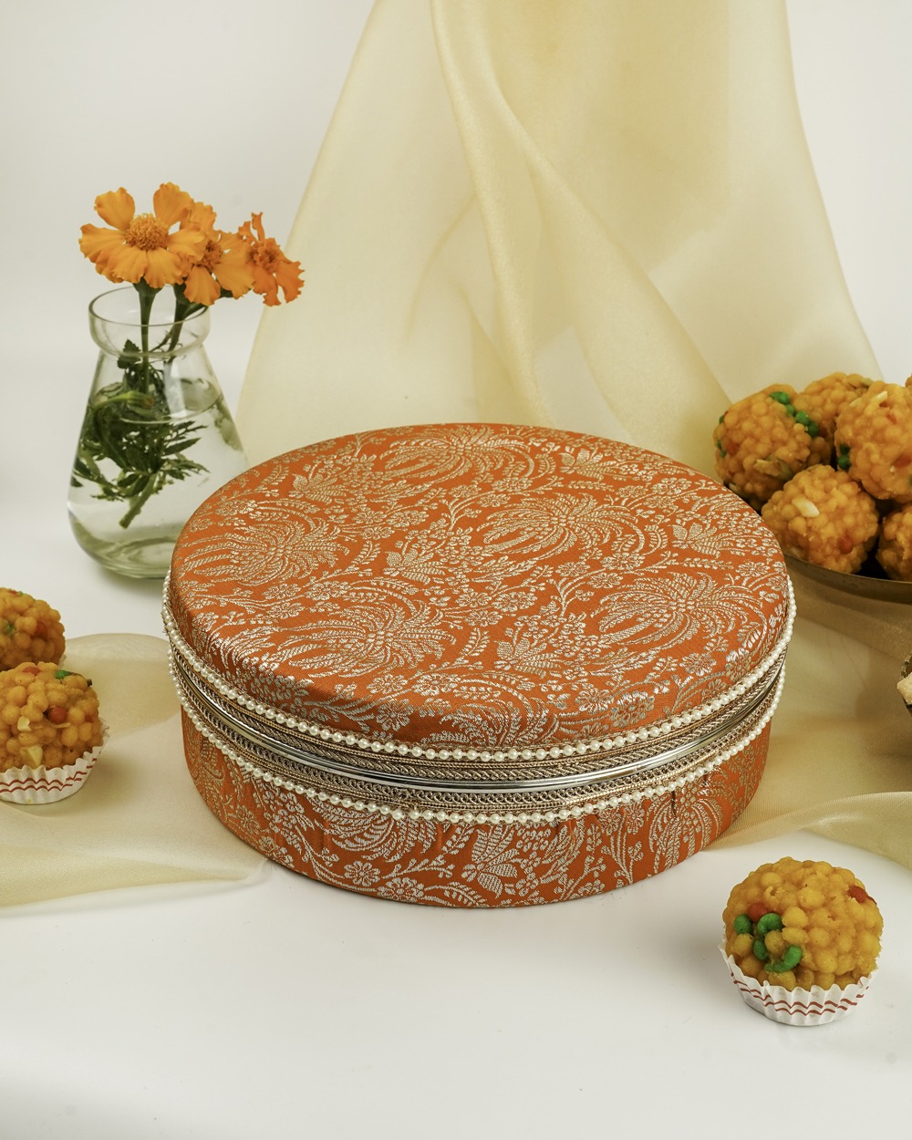 Vibrant orange Sunehri Ladoo Box by Navka Studio with white paisley brocade, silver lace, and a pearl border.