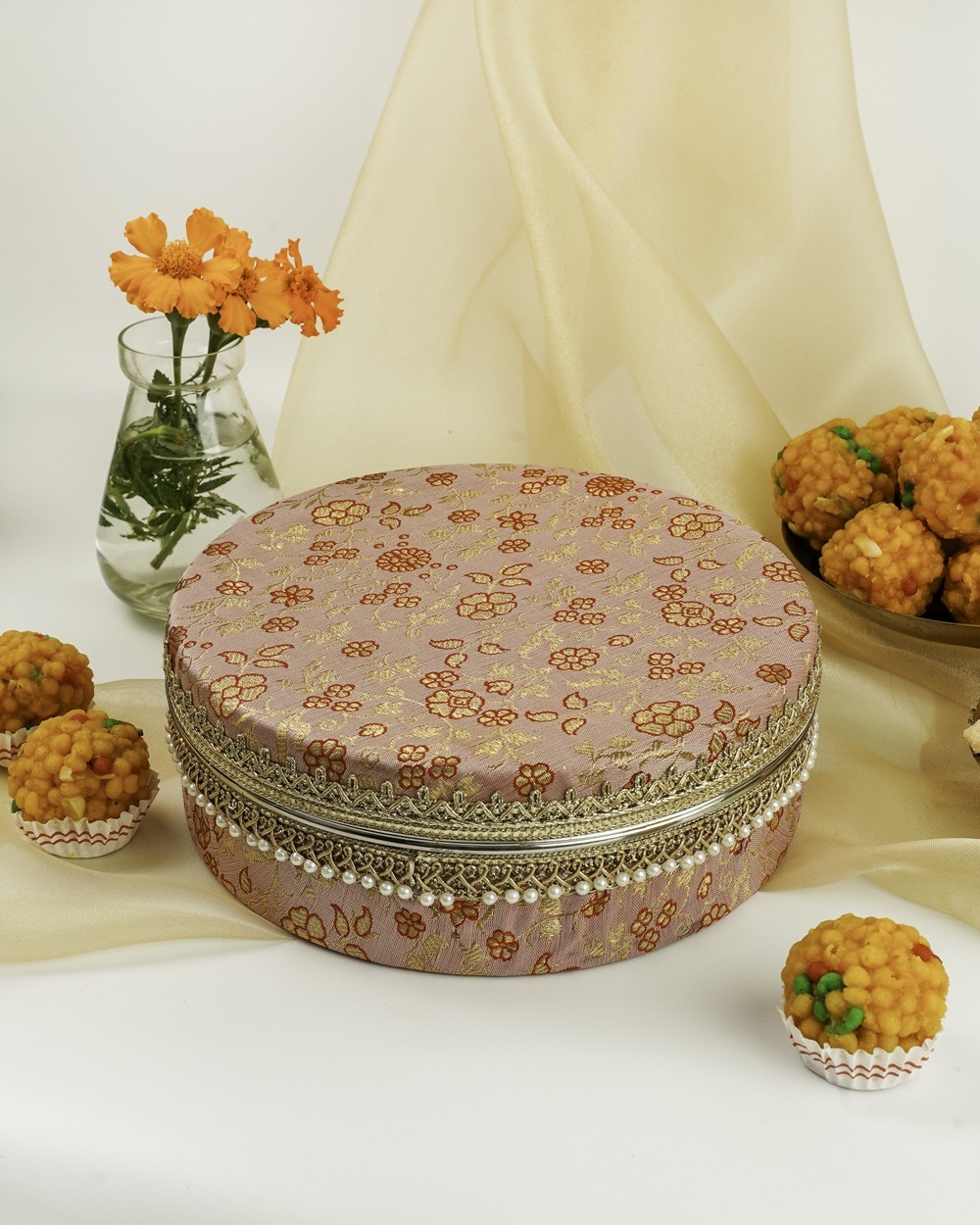 A charming dusty pink Bahaar Ladoo Box by Navka Studio, adorned with delicate gold and red floral embroidery, intricate gold lace, and a pearl-beaded trim, surrounded by colorful sweets and flowers.