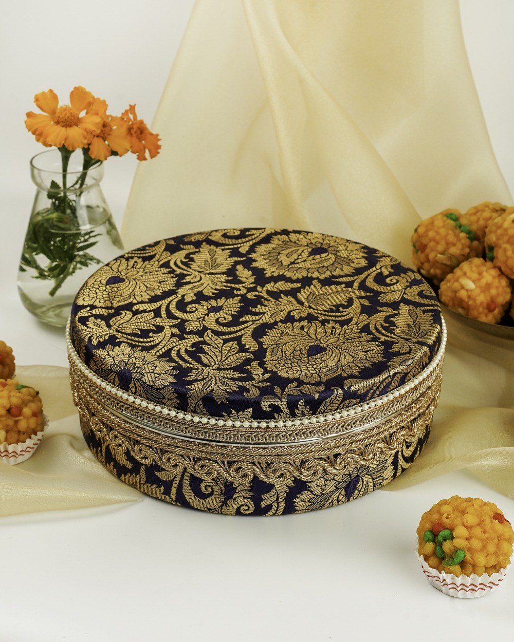 A regal navy blue Chandni Ladoo Box by Navka Studio, featuring intricate gold floral brocade, a wide gold and silver lace trim, and a delicate pearl-beaded border, displayed with festive sweets and marigold flowers.