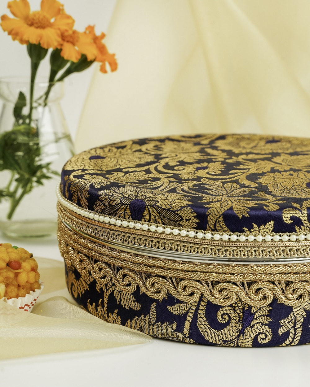 A regal Chandni Ladoo Box by Navka Studio, featuring a deep navy blue fabric lid with opulent gold floral and paisley patterns, adorned with elaborate gold braided trim and a delicate string of pearls along its edge. The box is subtly accompanied by a traditional Indian sweet (ladoo) in a red cup and a vase with vibrant orange flowers, set against a soft, draped background.