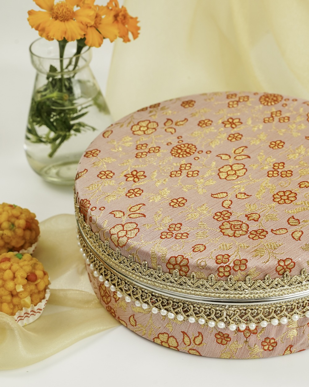 A close-up of the elegant dusty pink Bahaar Ladoo Box by Navka Studio, highlighting its delicate gold and red floral embroidery, intricate gold lace, and a border of small white pearls, with traditional sweets in the foreground.
