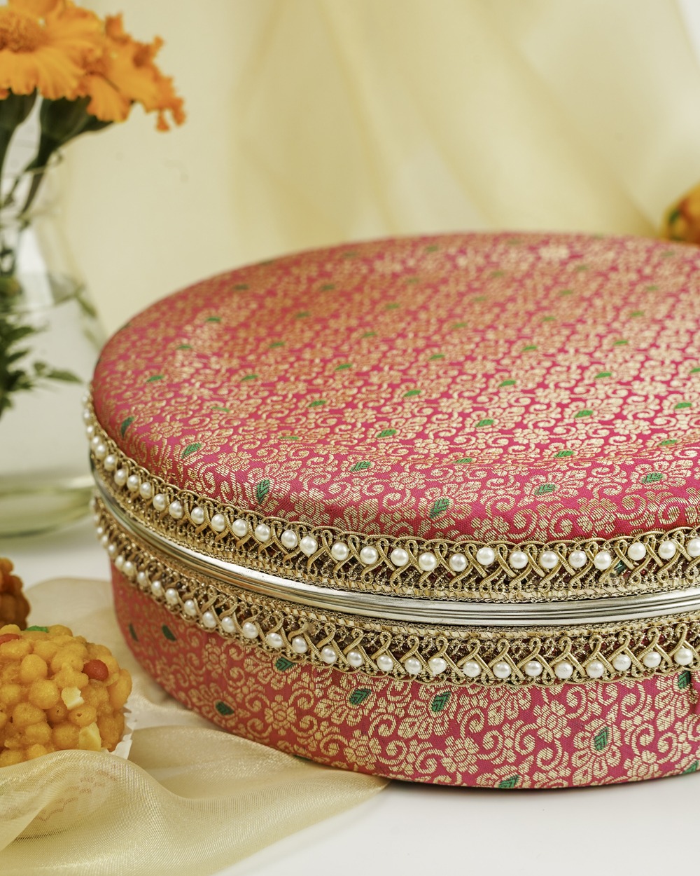 Close angle half view of Vibrant pink Gulzar Ladoo Box by Navka Studio with gold and green brocade, silver lace, and pearl trim, displayed with sweets.