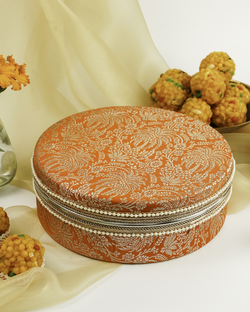Sunehri Ladoo Box for giftings in different angle Vibrant orange Sunehri Ladoo Box by Navka Studio with white paisley brocade, silver lace, and a pearl border.