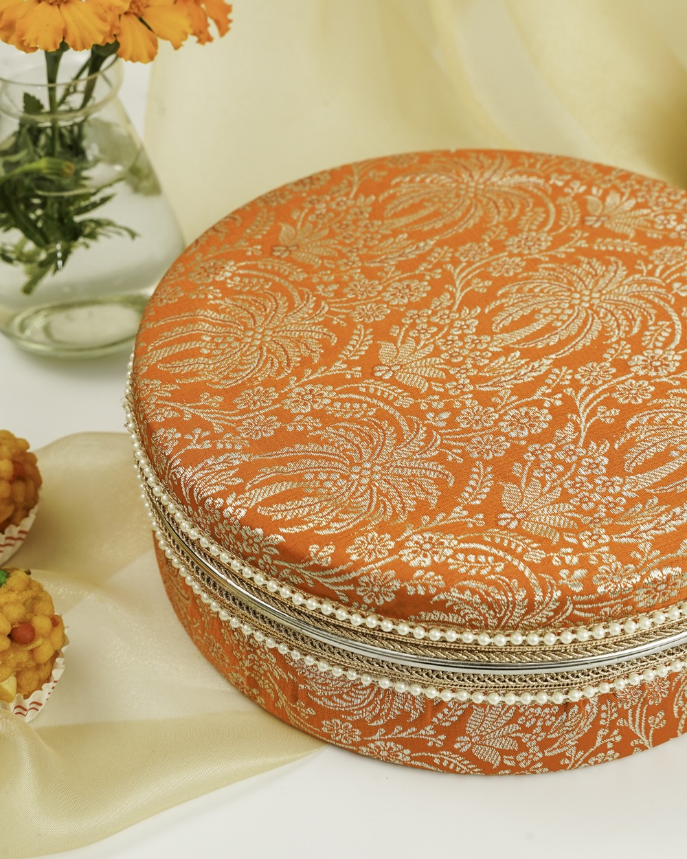 A vibrant orange Sunehri Ladoo Box by Navka Studio, featuring silver floral patterns, pearls, and delicate trim.