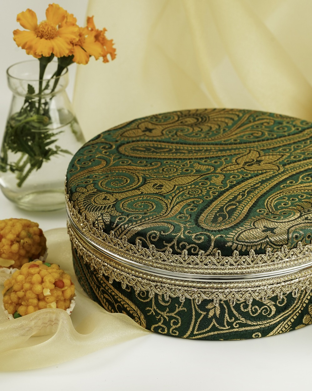 Navka Studio's round Baagh Ladoo Box, made from rich green fabric with shimmering gold paisley embroidery and a delicate silver lace border, displayed alongside traditional Indian ladoos.