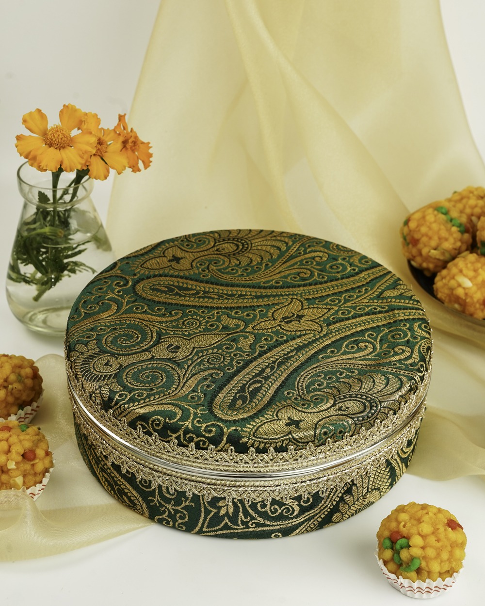 An opulent green and gold brocade Baagh Ladoo Box from Navka Studio, showcasing its detailed gold paisley pattern and silver-toned lace trim, presented in a festive setting with sweets.