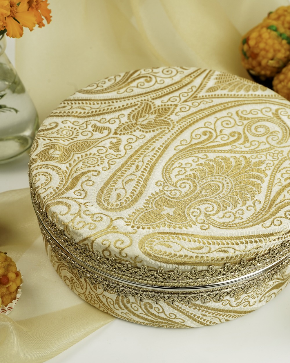 A detailed view of Navka Studio's Noori Ladoo Box, showcasing its cream and gold paisley brocade with gold lace trim.