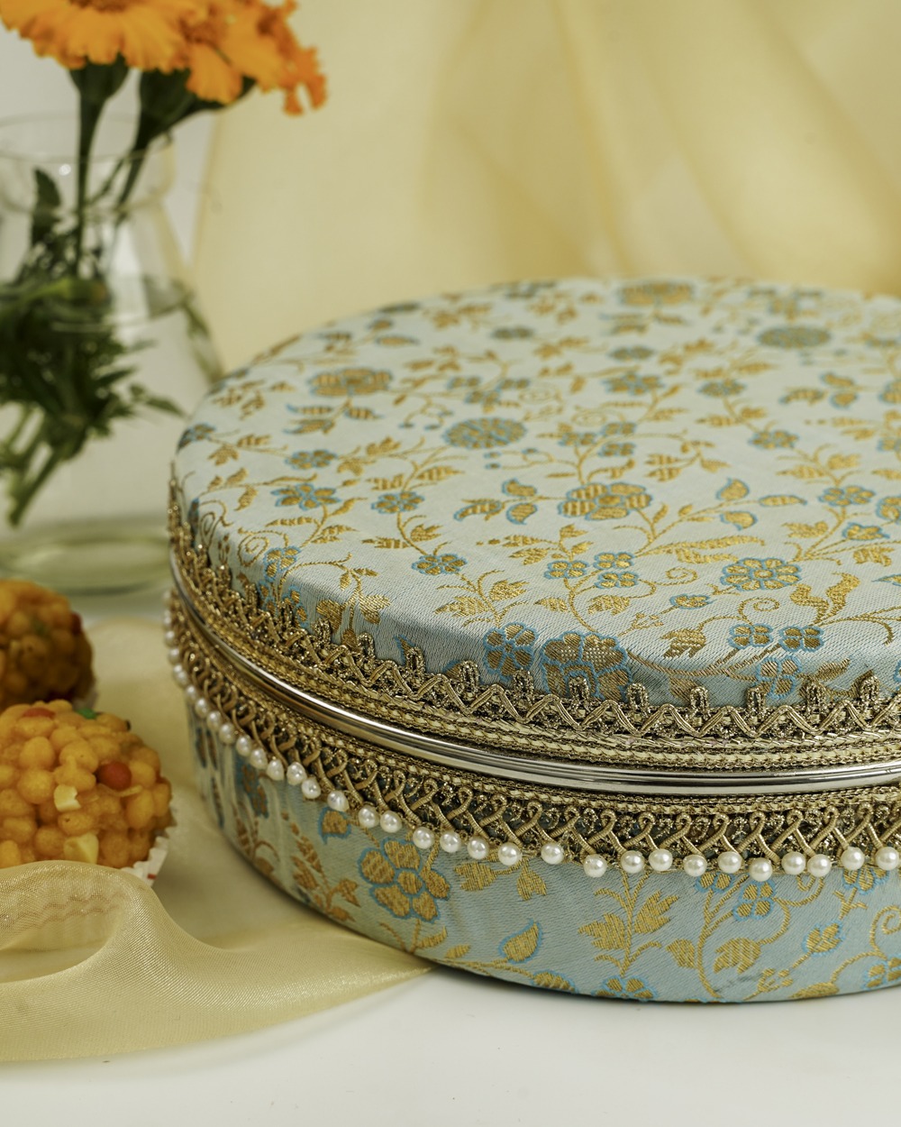 An exquisite light blue and gold embroidered Seher Ladoo Box from Navka Studio, showcasing its detailed floral design, golden lace, and pearl embellishments, presented in a festive setting with Indian sweets.