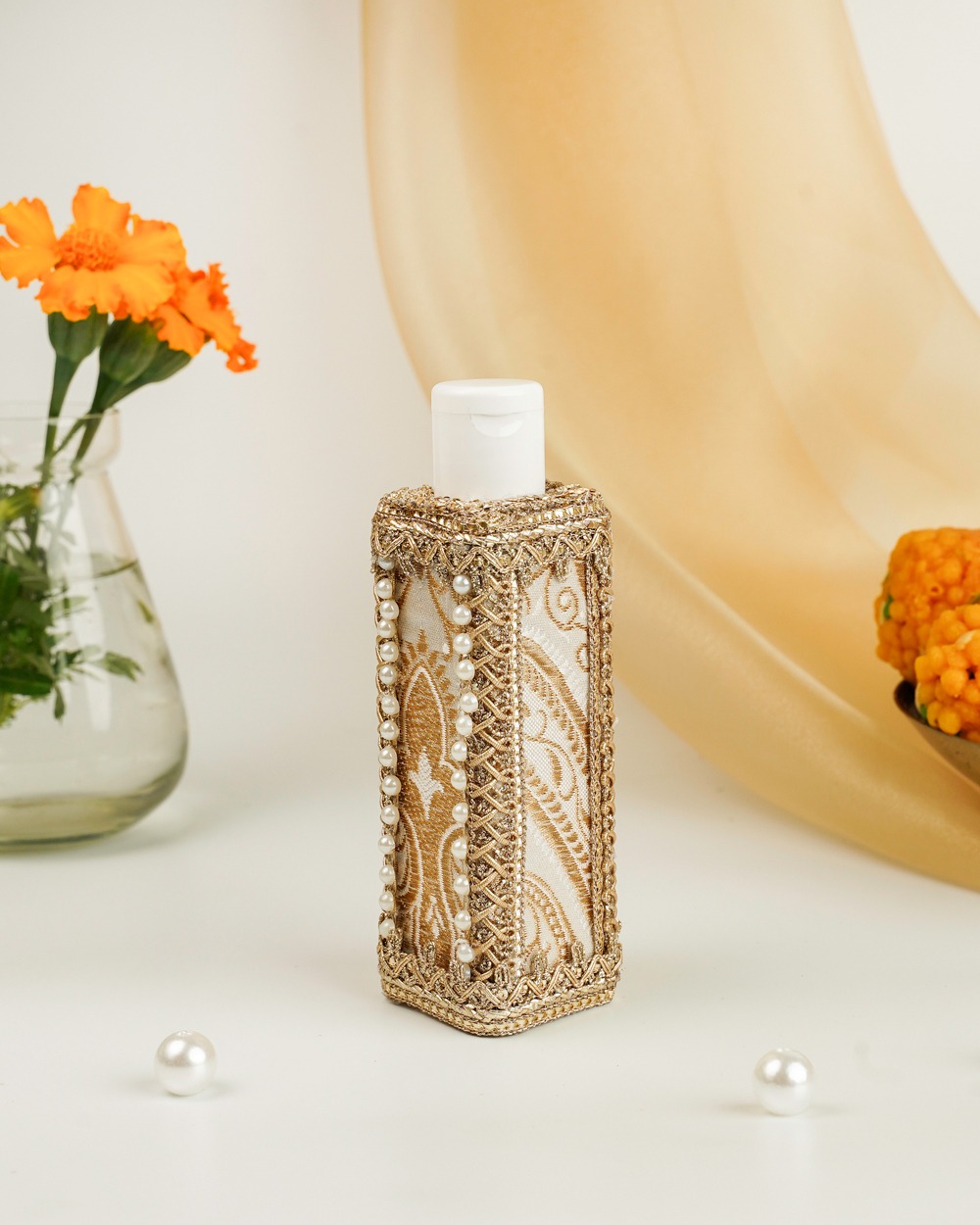 Noori Oil Bottle by Navka Studio – a decorative oil bottle with intricate golden embroidery, pearl accents, and elegant detailing, displayed with marigold flowers in the background.