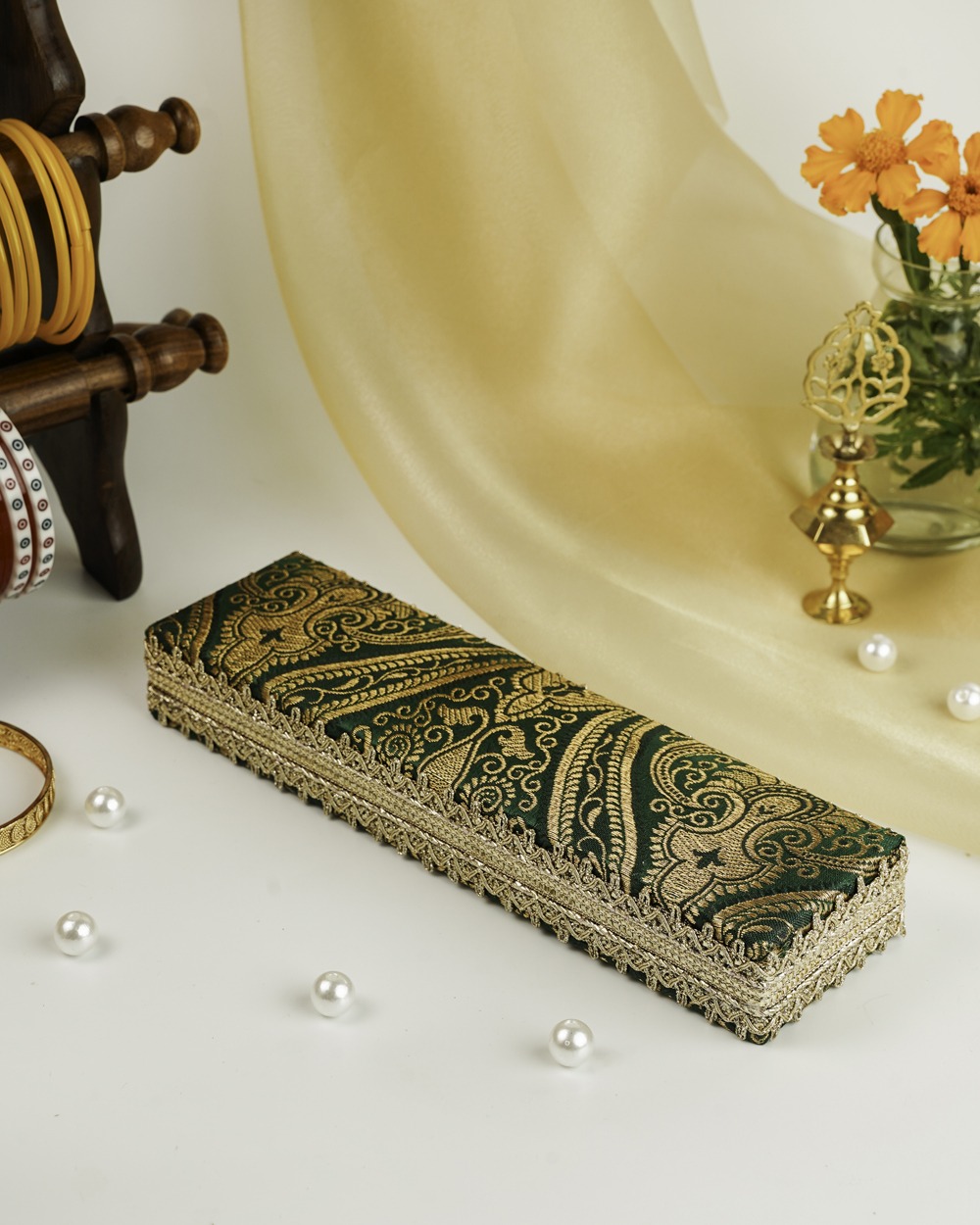 A Baagh Brocade Chain Box by Navka Studio in green and gold paisley fabric, with ornate lace trim, on a festive table.