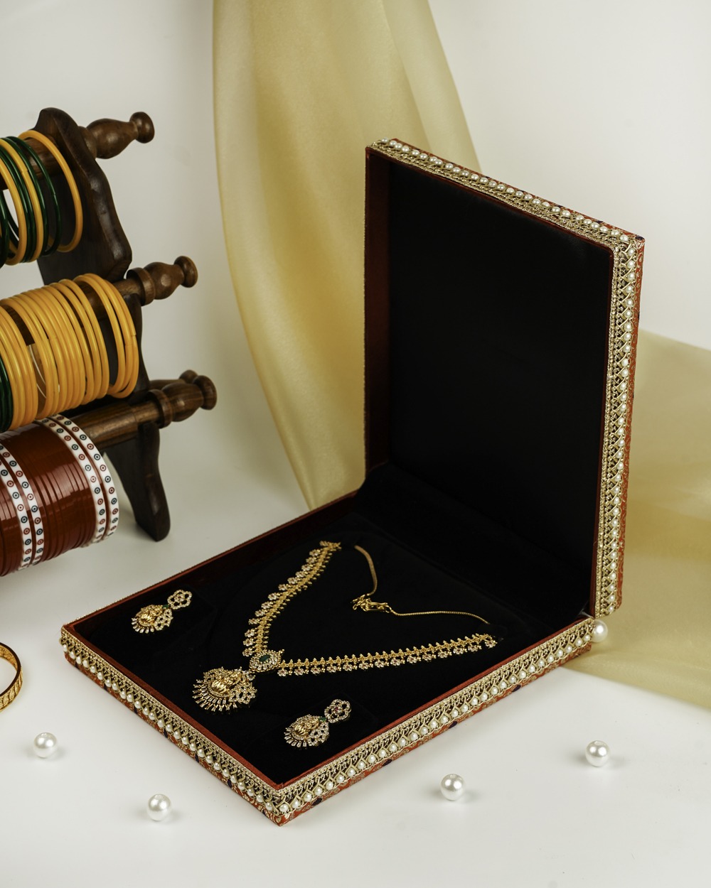 The Sindoori Brocade Necklace Box by Navka Studio, shown closed to highlight its vibrant red and gold brocade fabric and elegant pearl lace border, set against a festive backdrop.