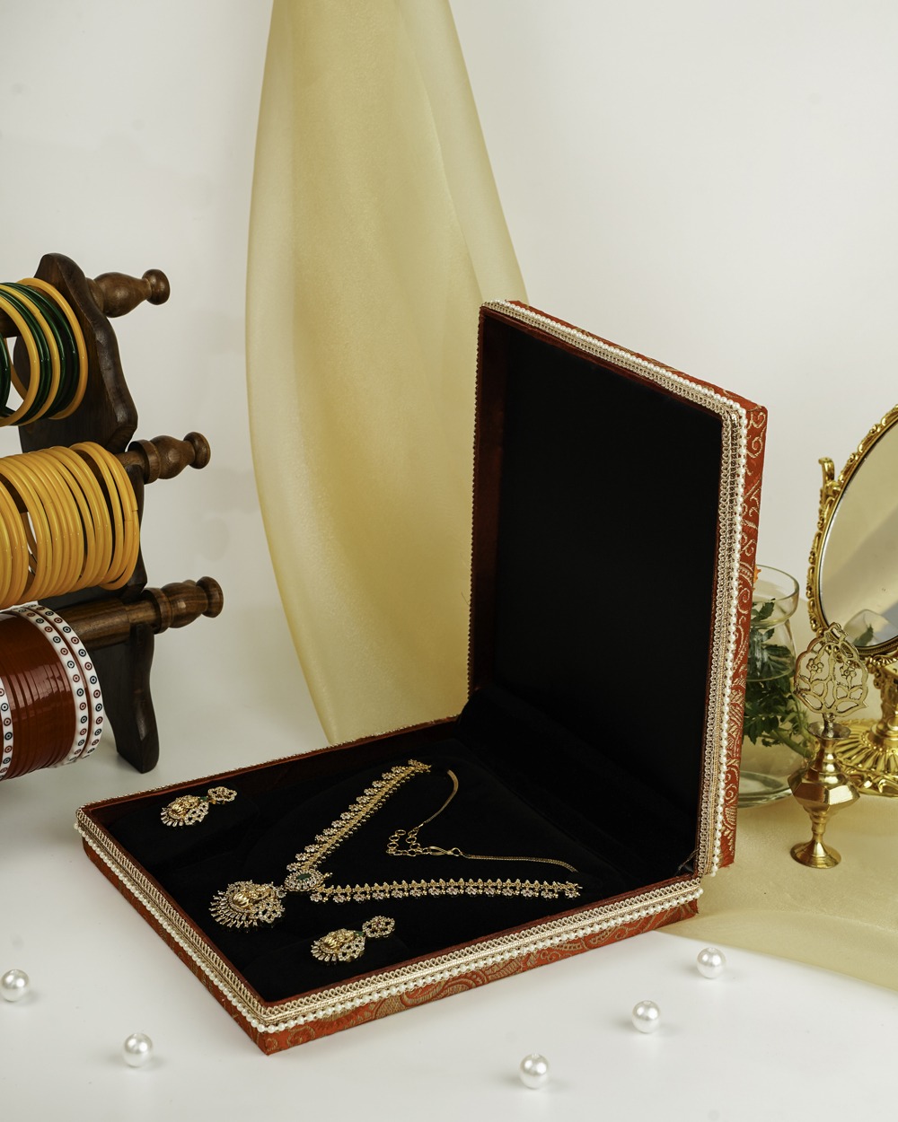 The Sindoori Paisley Brocade Necklace Box by Navka Studio, displayed open to showcase its black velvet interior holding a gold necklace set, placed next to a traditional bangle stand.