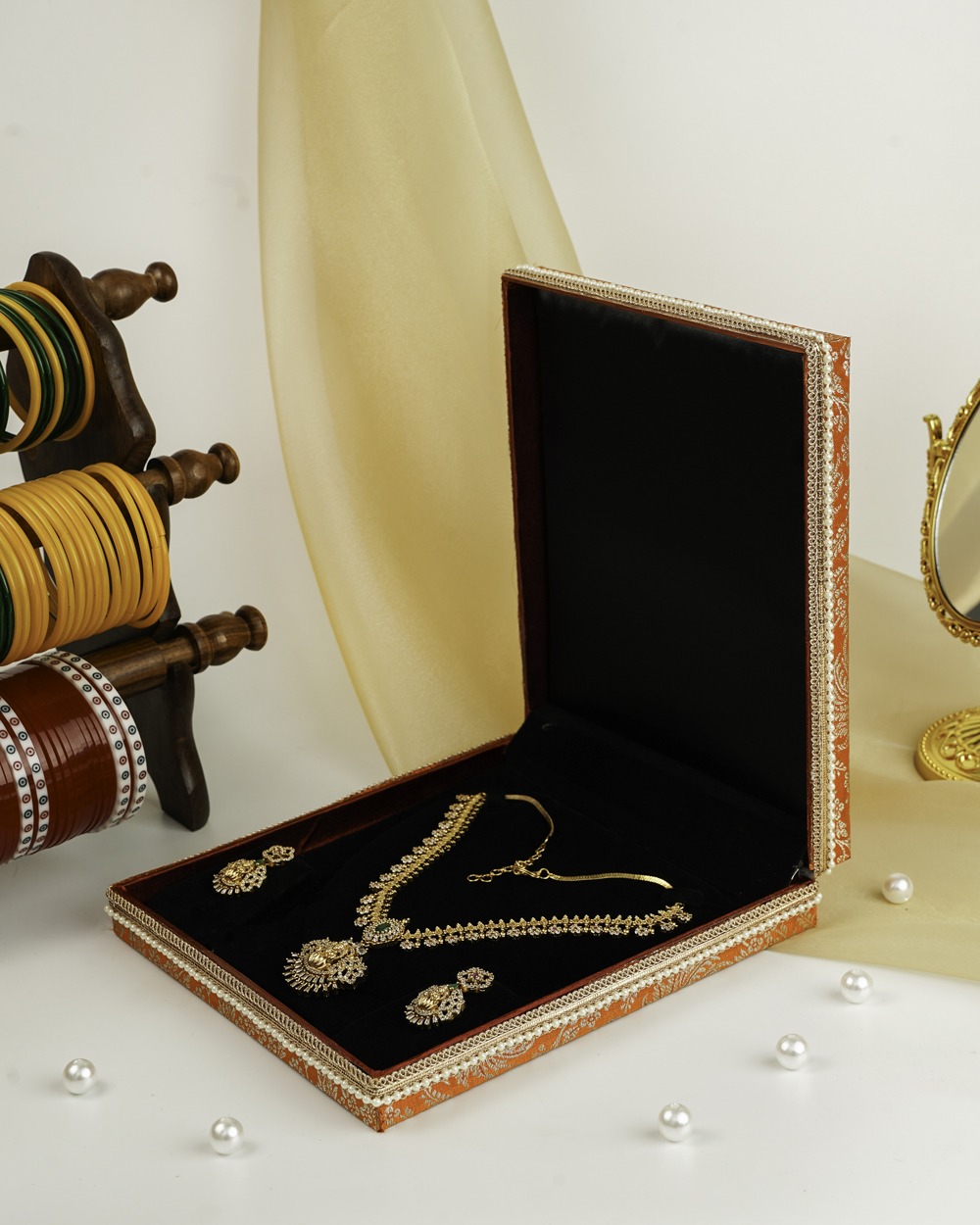 An orange Sunehri Brocade Necklace Box by Navka Studio, open, revealing a gold necklace and earring set on its black velvet lining.