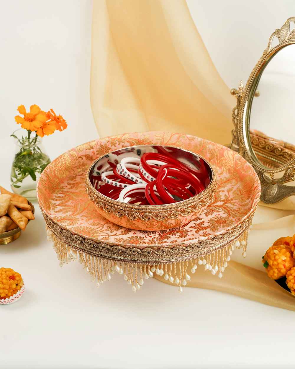 A Meher Chura Bowl by Navka Studio, filled with bangles, on its matching peach brocade tray with an elegant pearl fringe, set on a festive table.