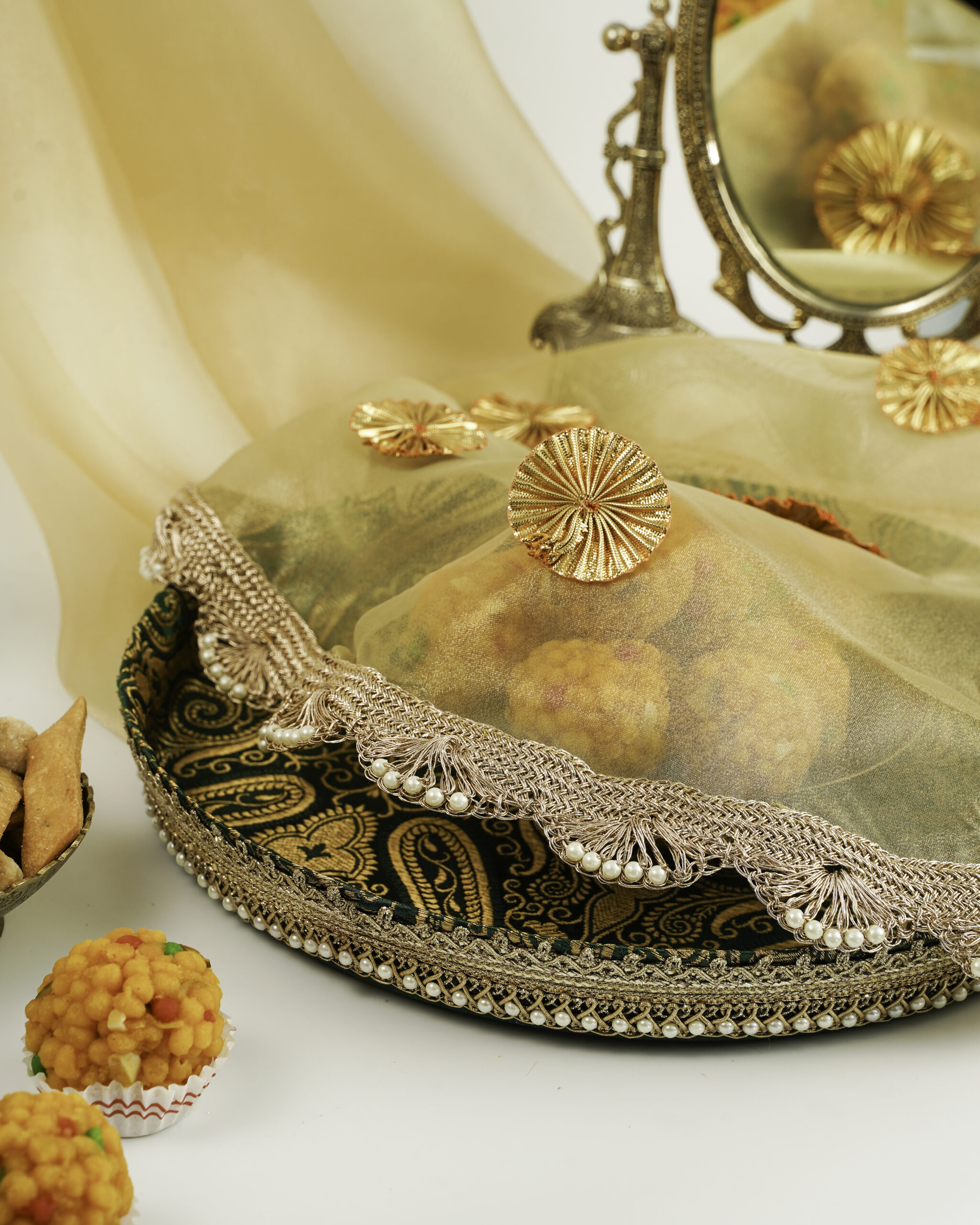 Elegant Baagh Shagun Thaal with Cover An elegant Baagh Shagun Thaal with Cover by Navka Studio, featuring green paisley brocade and a pearl border, shown with its sheer, lace-trimmed cover over ladoos.