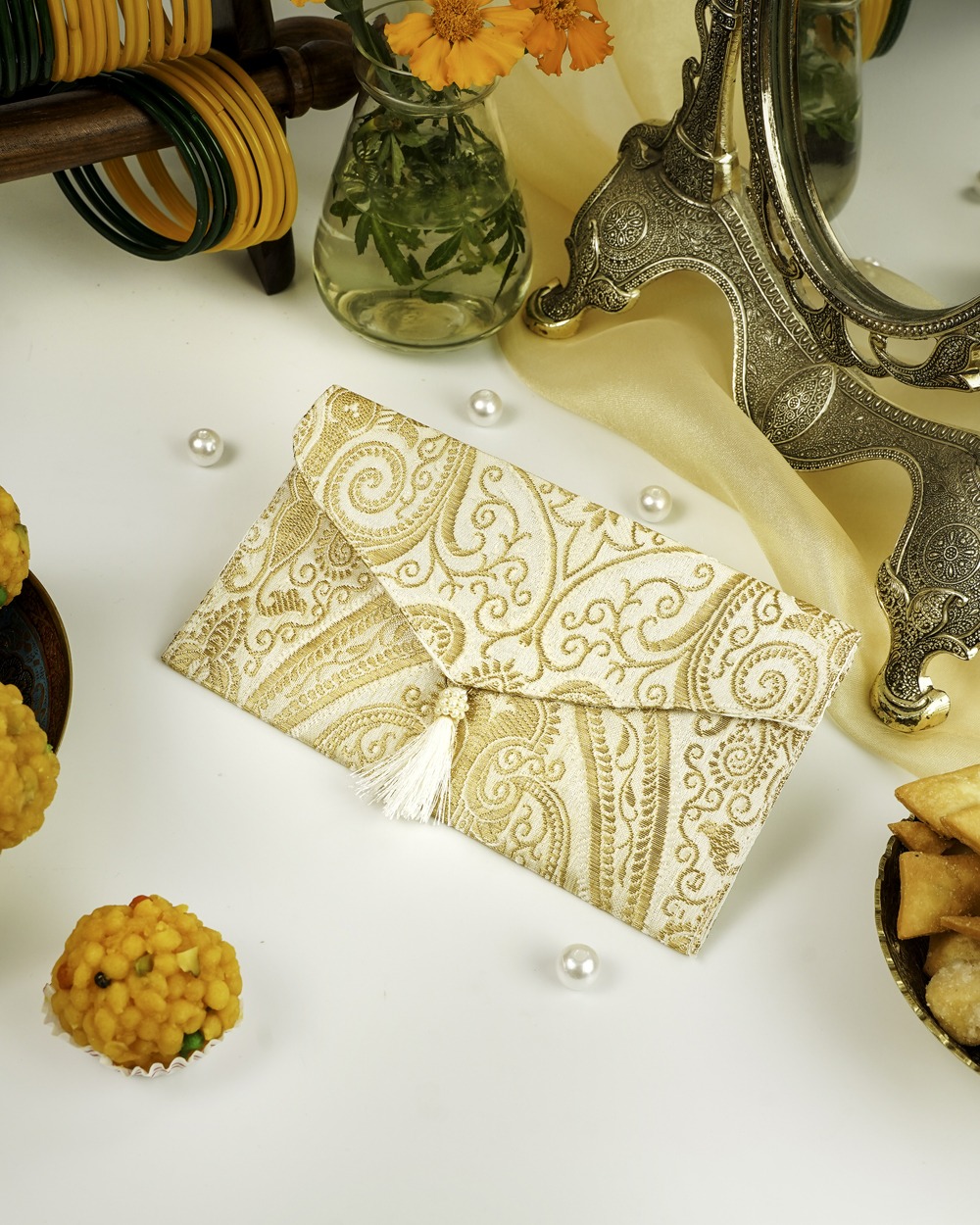 An elegant Noori Brocade Shagun Envelope by Navka Studio, in cream and gold paisley brocade with a white tassel, on a festive table.