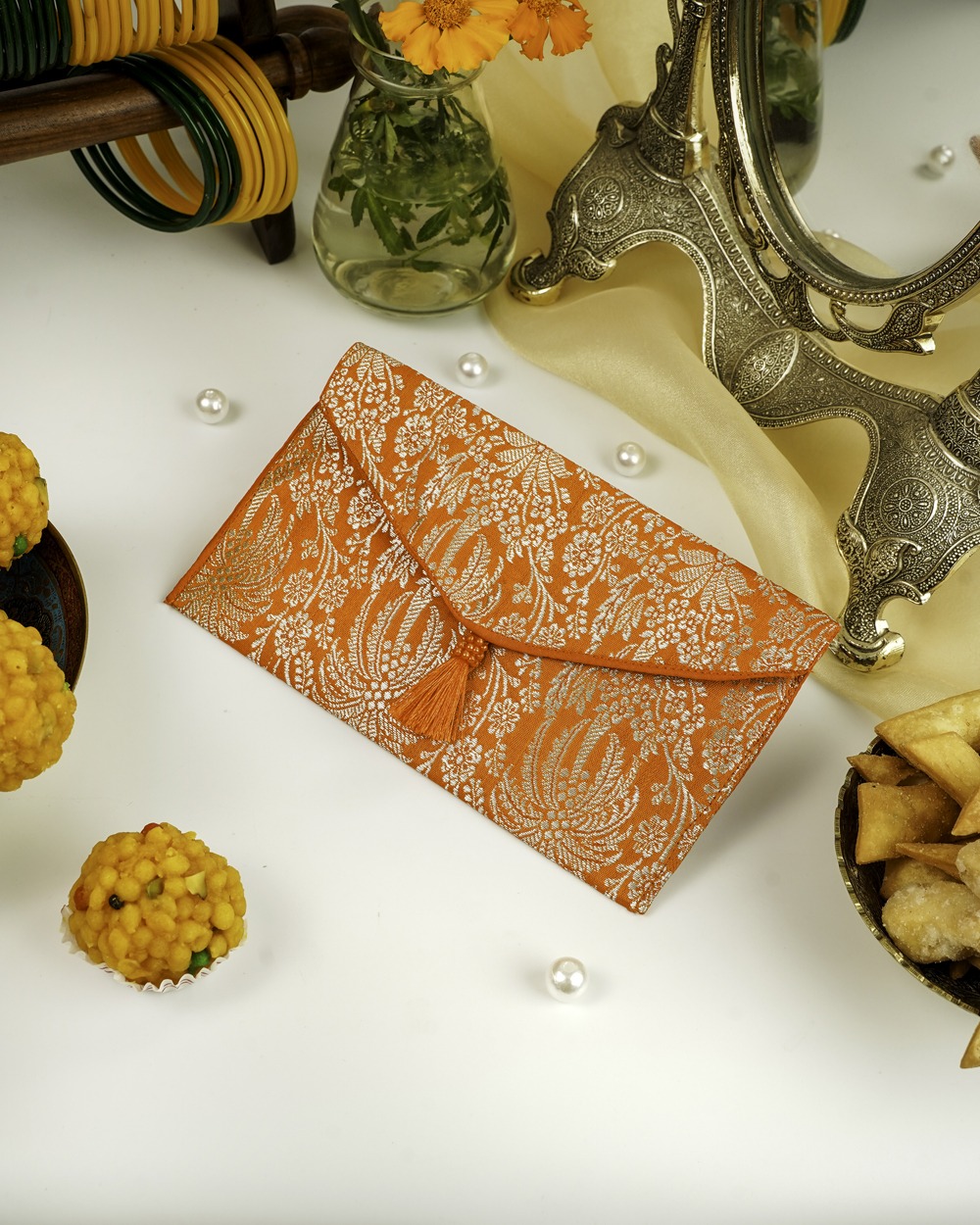 An orange Sunehri Brocade Shagun Envelope by Navka Studio, with silver floral brocade and a tassel, on a festive table.