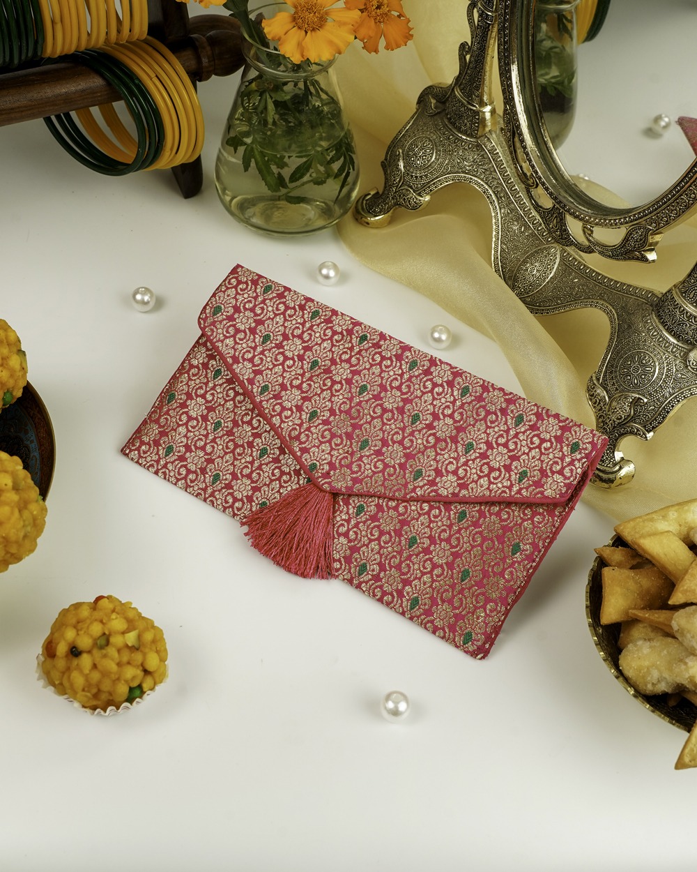 The Gulzar Brocade Shagun Envelope by Navka Studio, shown closed to highlight its vibrant pink and gold brocade fabric and a decorative pink tassel, resting on a festive surface.