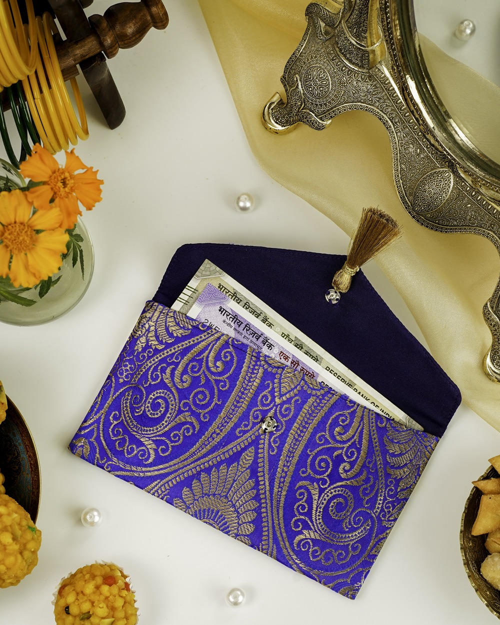 The Neelam Brocade Shagun Envelope by Navka Studio, shown closed to highlight its rich royal blue and gold paisley brocade fabric and a decorative gold tassel, placed on a festive table with sweets.