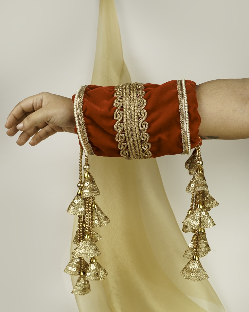 A red Sindoori Velvet Chura Cover - Big Tassel by Navka Studio, worn on an arm, with rich gold lace and large sequin tassels.