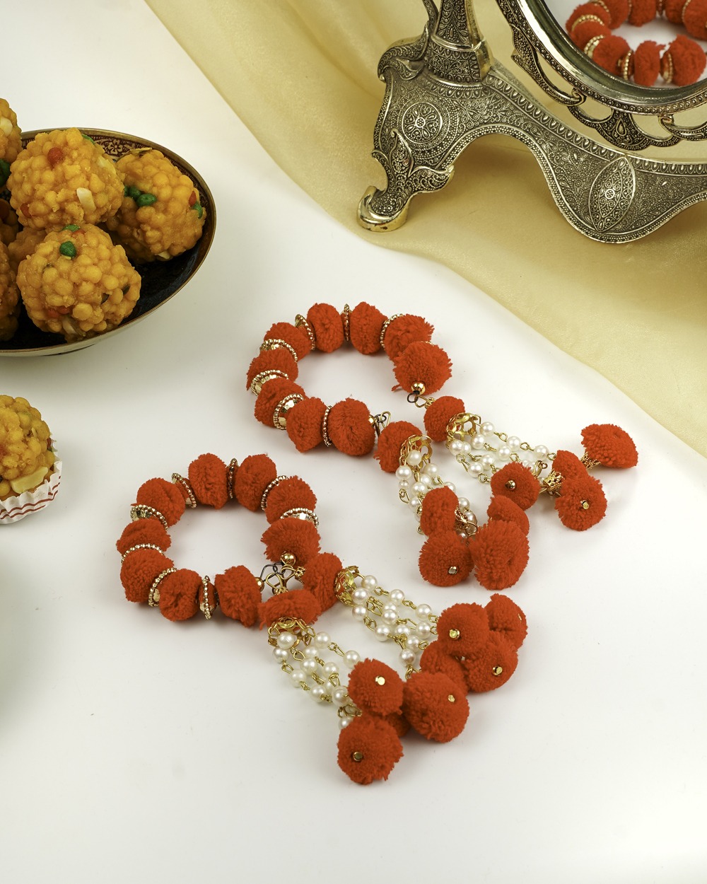 Two vibrant red pom-pom Sindoori Gaane by Navka Studio, adorned with gold accents and pearl strings, next to ladoos.