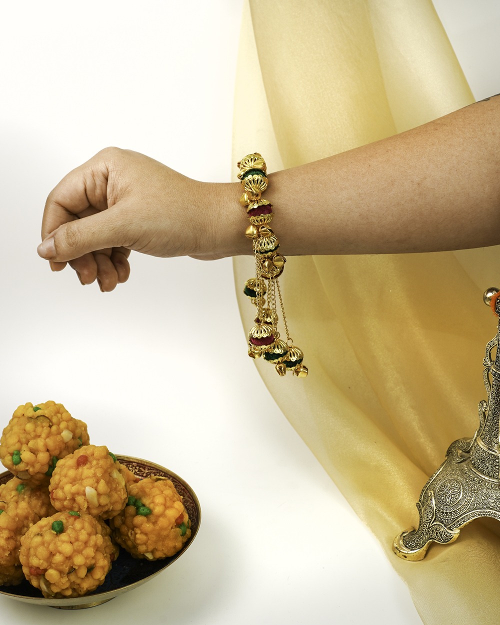A hand wearing a festive Sunehre Gaane by Navka Studio with colorful jhumki bells and gold accents, near ladoos.