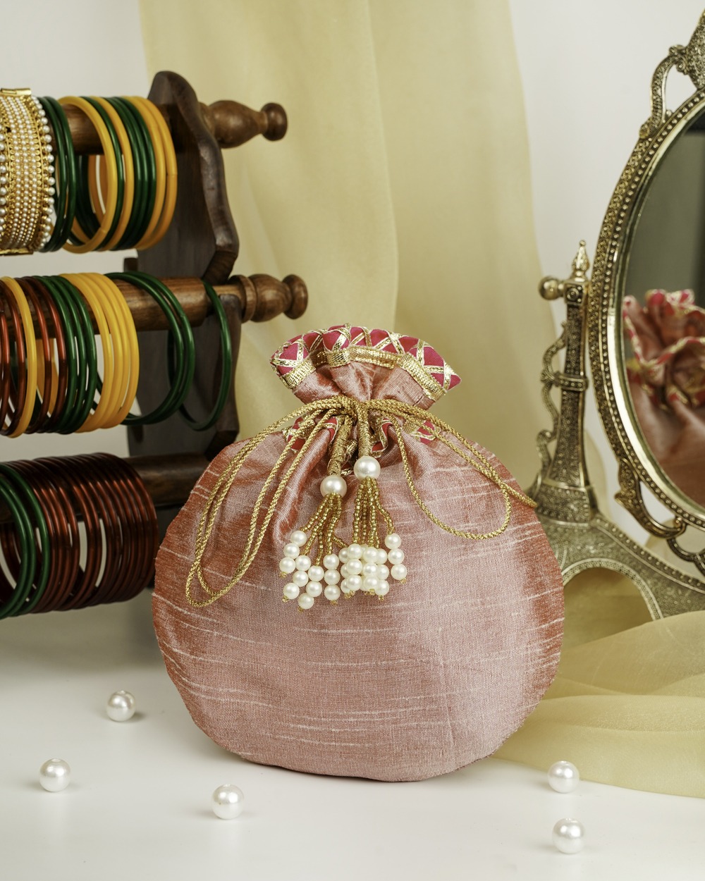 Navka's elegant Bahaar Silk Potli in a shimmering dusty pink, featuring a vibrant fuchsia and gold trim with delicate pearl tassels, styled on a vanity table.