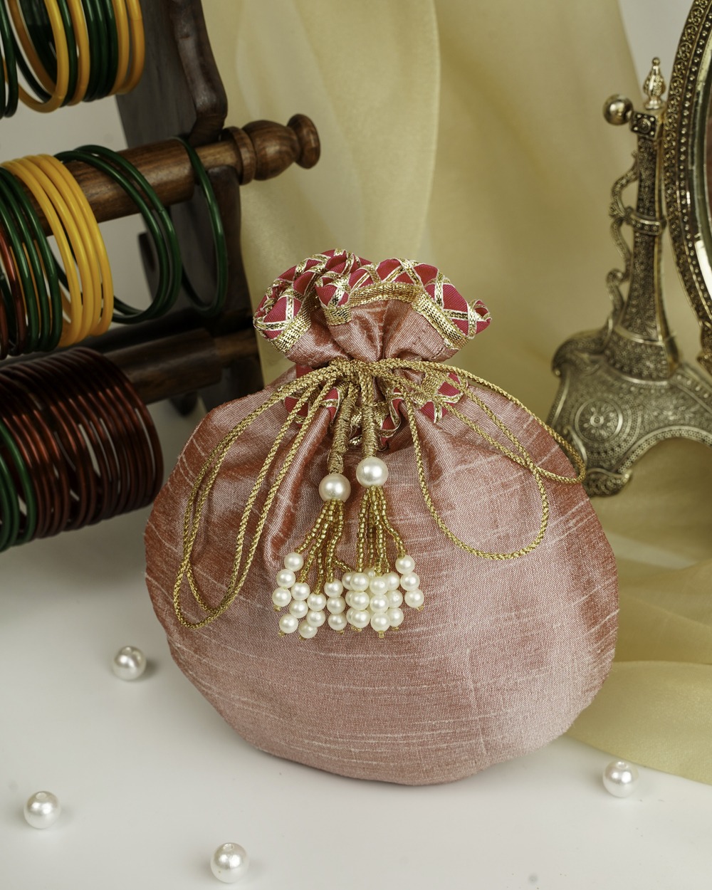 An elegant Bahaar Silk Potli by Navka Studio in shimmering dusty pink, featuring a pink and gold embroidered opening and beautiful pearl tassels, displayed on a dresser.