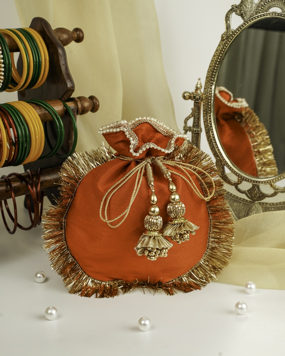 A vibrant orange Sunehri Silk Potli by Navka Studio, featuring a lush golden fringe border, a decorative red and gold patterned opening, and ornate golden bell tassels, displayed on a white surface.