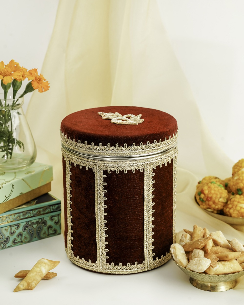 A luxurious red Saaj Velvet Ladoo/Bhaji Box by Navka, adorned with intricate lace, sitting on a festive table next to a bowl of Indian sweets.