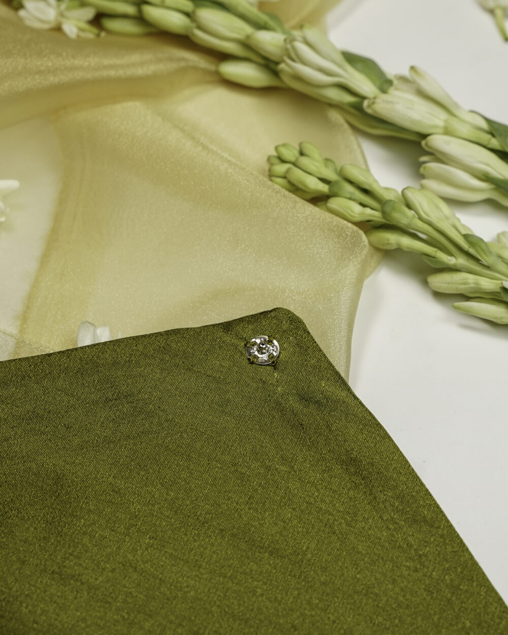 Close-up of Saawan Silk, Full Gota work with Gold trims, Trousseau Bag, an olive green fabric envelope with a small silver floral button, placed on a golden cloth with white flowers in the background by Navka Studio.