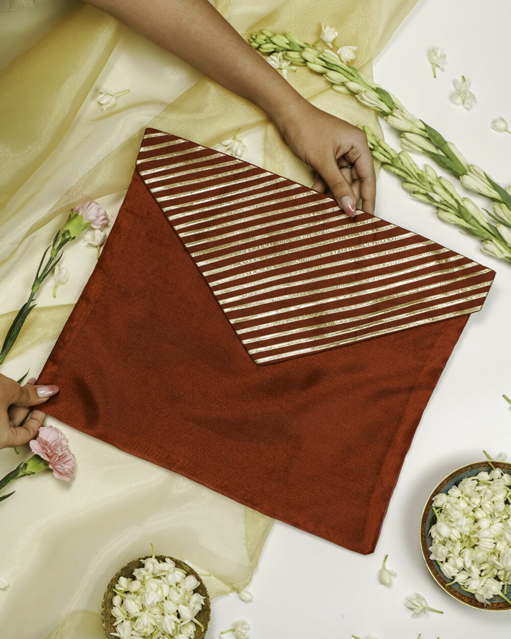 a person holding Saaj Silk, Full Gota work, Trousseau Bag, a red and gold striped fabric envelope or clutch, placed on a white surface with white flowers and a golden cloth in the background by Navka Studio.