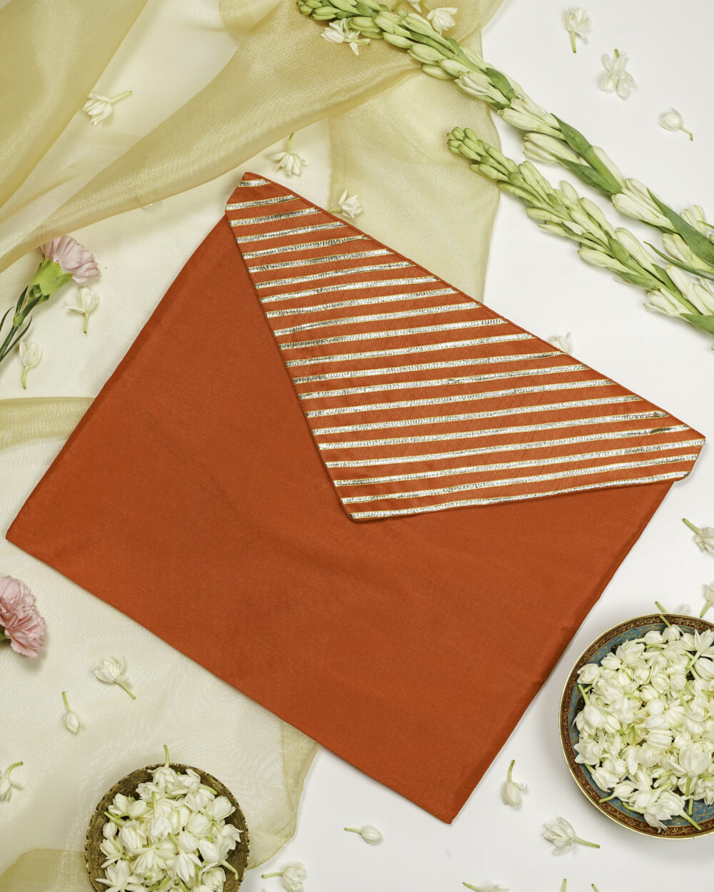 Suneheri Silk, Full Gota work, Trousseau Bag, a bright red envelope-style fabric pouch with a flap featuring gold and red horizontal stripes, placed on a white surface decorated with white flowers, gold fabric, and flower bowls By Navka Studio