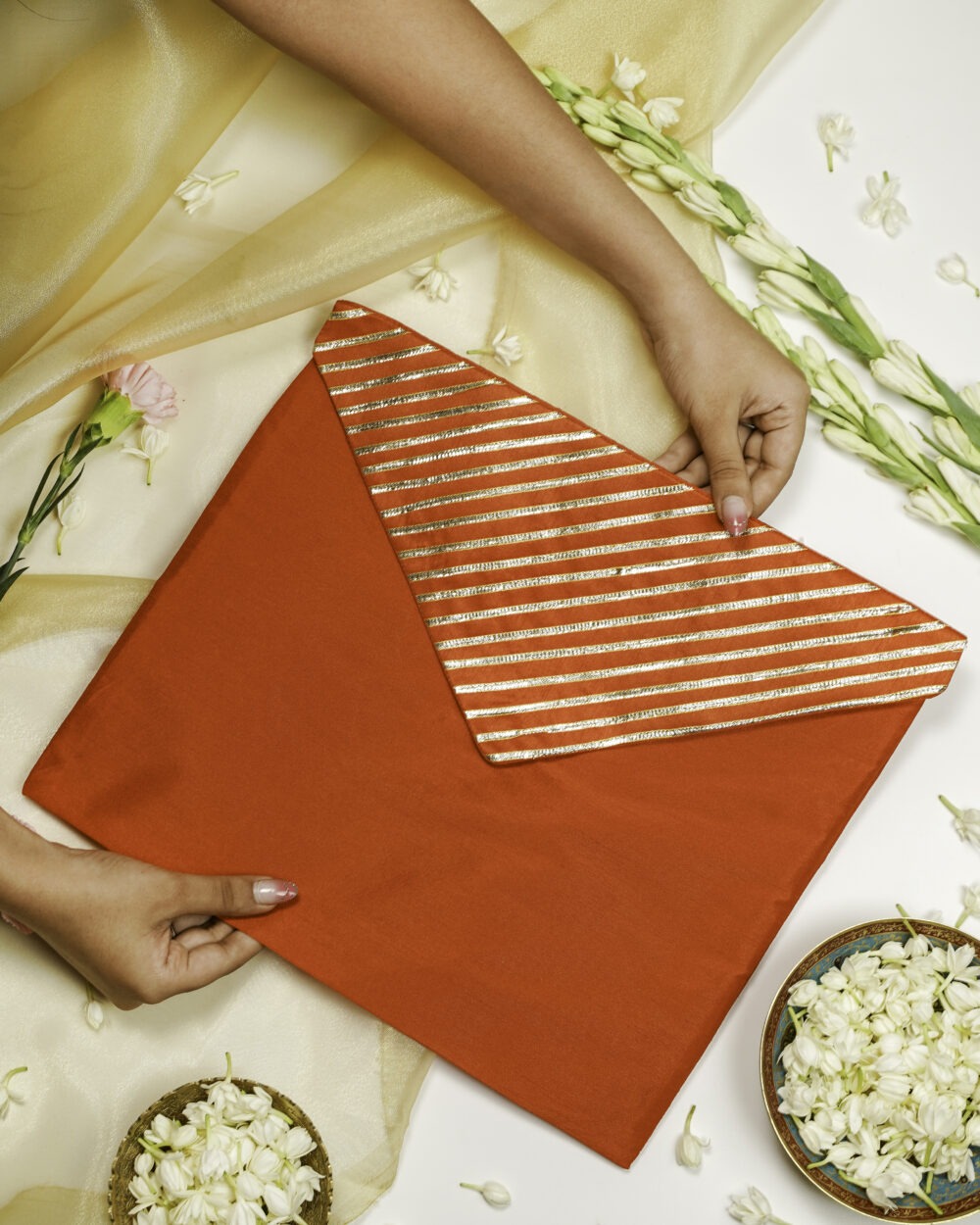 A person holding Suneheri Silk, Full Gota work, Trousseau Bag, a bright red envelope-style fabric pouch with a flap featuring gold and red horizontal stripes, placed on a white surface decorated with white flowers, gold fabric, and flower bowls.