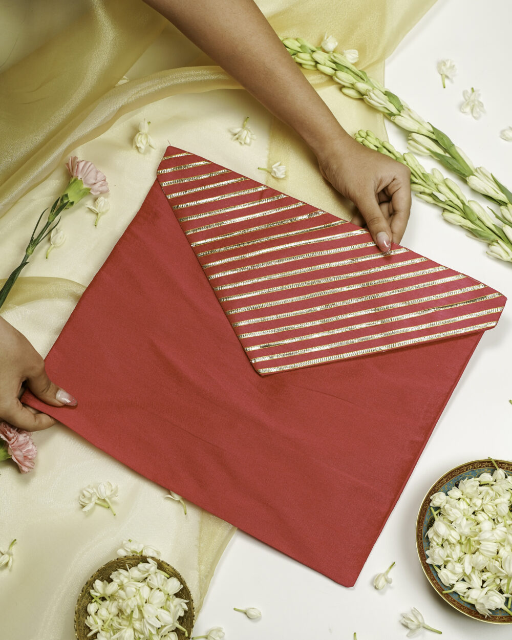 Navka Studio Gulzaar Silk, Full Gota Work, Trousseau Bag — a red silk envelope-style bag with a golden striped gota flap, held open by hands over a floral background.