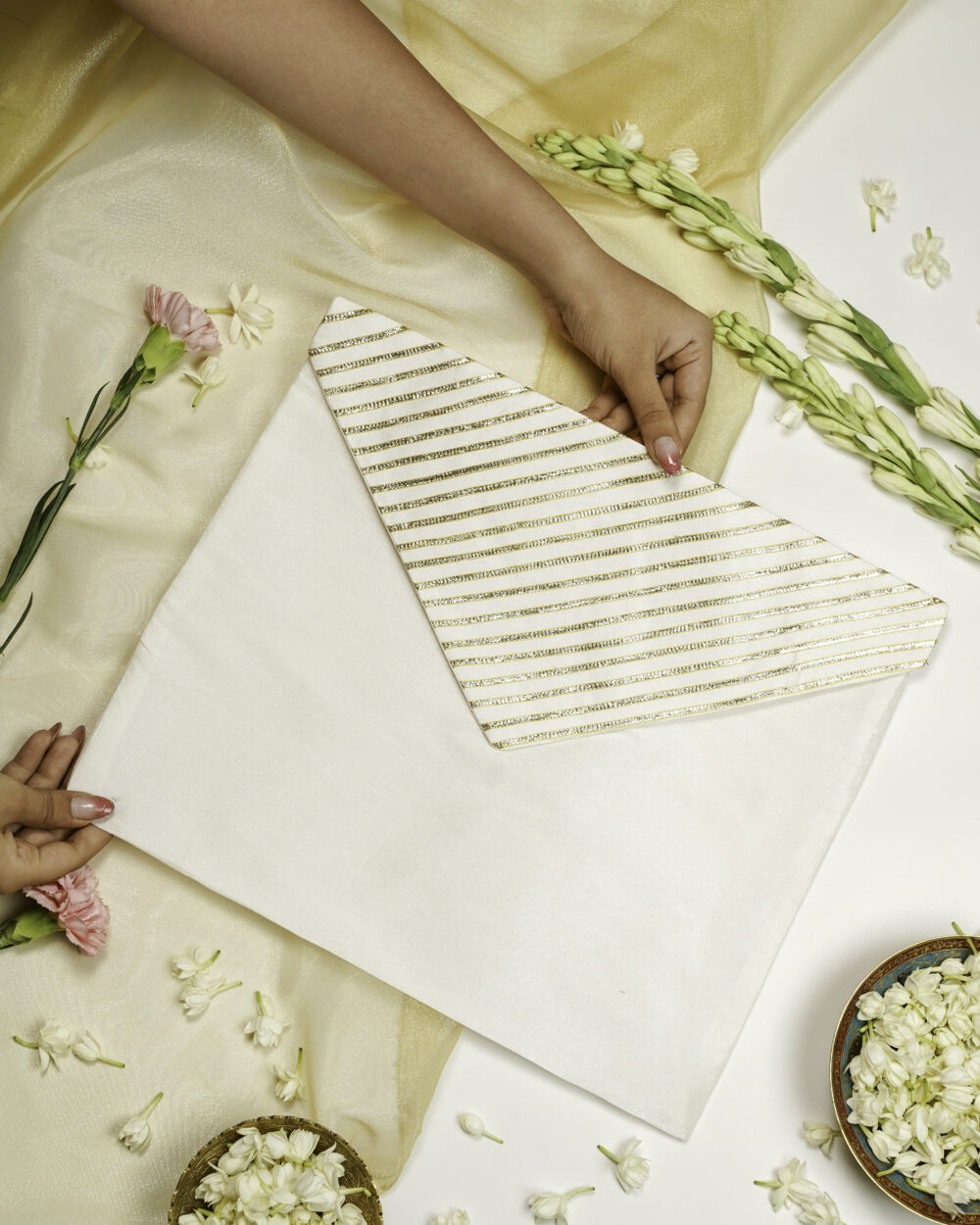 Two hands holding Noori Silk, Full Gota work with Gold trims, Trousseau Bag, an elegant white fabric envelope with a flap featuring diagonal gold stripes, placed on a white surface adorned with golden cloth, flowers, and bowls filled with white petals by Navka Studio.