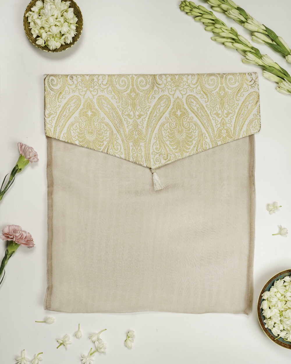 Noori Brocade Silk & Organza Trousseau Bag, A beige fabric envelope with a flap featuring an intricate gold paisley design and a small tassel, placed on a white surface decorated with flowers and bowls of white petals by Navka Studio.