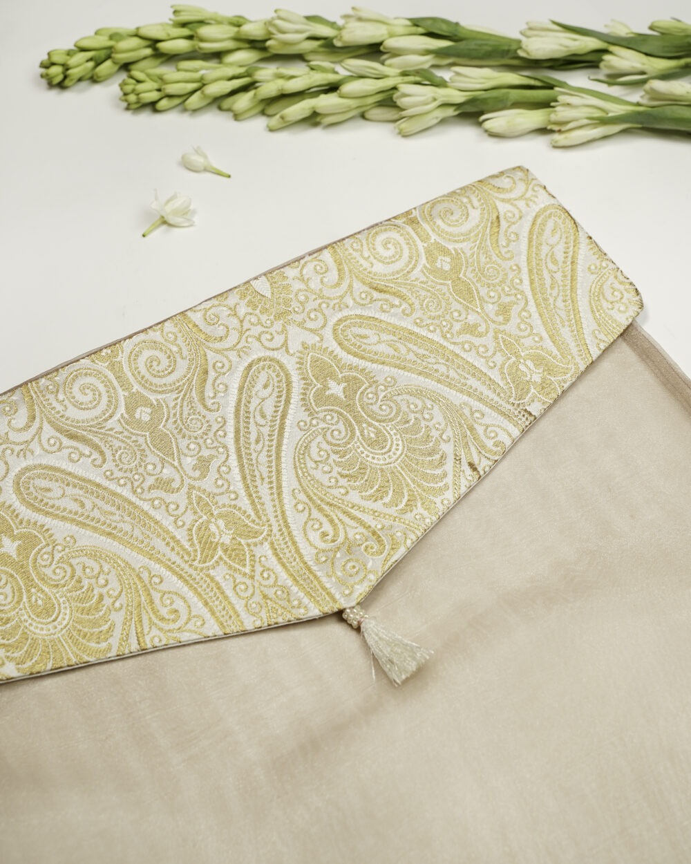Noori Brocade Silk & Organza Trousseau Bag, A beige fabric envelope with a flap featuring an intricate gold paisley design and a small tassel, placed on a white surface decorated with flowers and bowls of white petals Navka Studio.