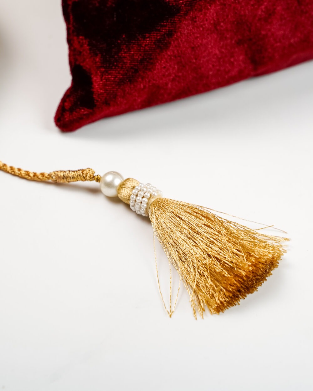 Saaj Silk Potli ,Golden tassel with pearl and bead detailing placed beside a red velvet fabric.